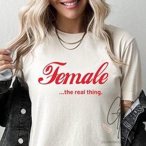 May include: A cream-coloured t-shirt featuring the word "Female" in red script, with the text "...the real thing" underneath. The shirt is a casual style. The image is a close-up of the t-shirt.
