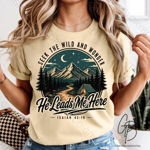 May include: A pale yellow t-shirt with a circular graphic. The graphic includes the text "Seek the Wild and Wonder" and "He Leads Me Here." It depicts a mountain scene with a tent, trees, and a crescent moon. The Bible verse Isaiah 43:19 is also included.