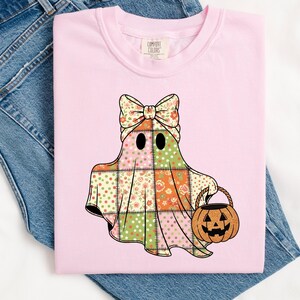 May include: A light pink Comfort Colors t-shirt featuring a patchwork ghost design. The ghost has a floral bow, black eyes, and a patchwork body in pink, green, and orange. It carries a jack-o'-lantern basket, perfect for Halloween.