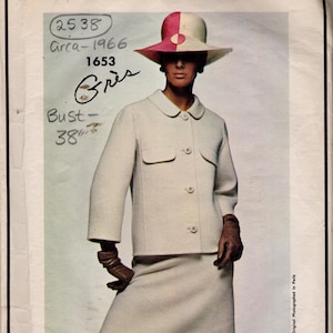 May include: A vintage Vogue Paris Original sewing pattern envelope featuring a cream-coloured jacket with a rounded collar, three-quarter sleeves, and two flap pockets, paired with a matching skirt, a two-toned pink and cream hat, and brown gloves.