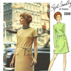May include: A vintage Vogue Couturier Design sewing pattern featuring a dress with a tie belt. The pattern is for a short-sleeved dress with a round neckline and pockets. The pattern number is 2359. The pattern is by Sybil Connolly of Dublin.