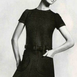 May include: A black, short-sleeved dress with a fitted shape. The dress has a belt at the waist, two front pockets, and a knee-length hemline. The model is wearing the dress and has her hand in one of the pockets.