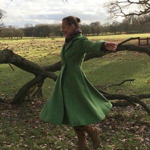 Puede incluir: Una mujer con un abrigo verde con falda acampanada, de pie en un campo con una rama de árbol detrás de ella. Lleva botas marrones y tiene los brazos extendidos.