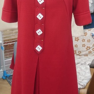 May include: A red dress with a white collar and white buttons down the front. The dress has short sleeves and a pleated skirt.