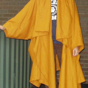 Puede incluir: Una larga capa de color amarillo mostaza con cuello ancho y estilo envolvente. La capa está hecha de una tela suave y fluida y tiene un corte holgado.