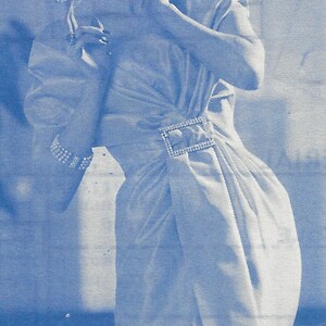 May include: A woman in a white dress with a beaded belt smiles and looks to the right. The dress has a cinched waist and a flowing skirt.