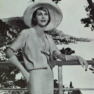 May include: A woman wearing a white hat and a light grey suit stands on a balcony overlooking a scenic view. She is wearing white gloves and has her hand resting on a railing.
