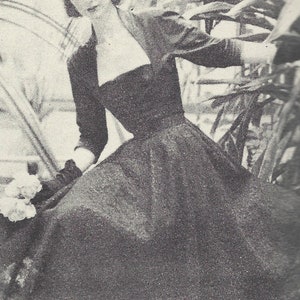 Peut inclure: Une robe noire avec un corsage ajusté et une jupe ample. La robe est portée par une femme avec les cheveux tirés en arrière. La femme se tient devant un fond feuillu.