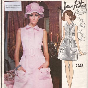 Op de afbeelding: Een vintage Vogue Paris Original naaipatroon-envelop met een mouwloze lichtroze jurk met kraag, een brede riem en twee voorzakken met knopen. De envelop bevat een bijpassende roze hoed met bloemaccenten en tekst.
