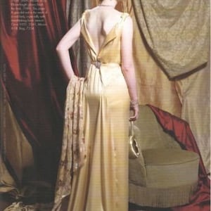 May include: A woman wearing a long, flowing, gold satin dress with a low back and a floral print scarf. She is standing in front of a red and gold patterned backdrop.