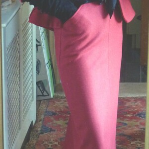 May include: A woman wearing a red suit with a black and red scarf. She is standing on a patterned rug and wearing red heels.