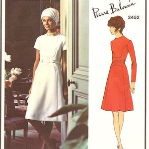 May include: Vogue Paris Original pattern number 2452 by Pierre Balmain. The pattern features two illustrations of women wearing dresses. The woman on the left is wearing a white dress with a belt and the woman on the right is wearing a red dress with a belt.