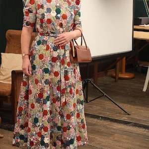 May include: A woman in a vintage-style dress with a colorful patchwork pattern. The dress has short sleeves, a button-down front, and a matching belt. She carries a brown handbag. The text "Dig for Victory - 1940s Fashion Show - June 2025" is at the bottom.