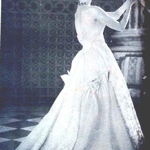 May include: A woman wearing a white gown with a large bow at the back. She is standing in front of a patterned wall and holding a piece of fabric.