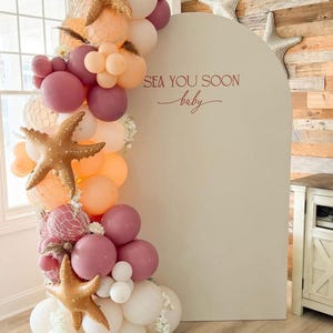 May include: A decorative balloon arch with a mix of peach, white, and mauve balloons, adorned with gold starfish and netting. The arch is set against a neutral backdrop with the text "Sea You Soon baby".