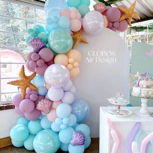 May include: A mermaid-themed balloon arch in pastel colors, including blue, pink, and purple. The arch features pearl-like balloons, seashell and starfish decorations, and the text "GLOBOS Air Design".