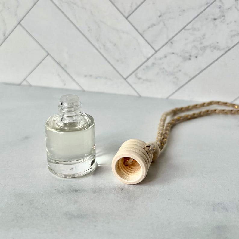 以下が含まれることがあります： 明るい色の液体が入った透明なガラス瓶と、天然ロープが付いた木製のディフューザー。 ディフューザーは、彫刻されたリングが付いた円筒形です。 白い表面に置かれたアイテム。