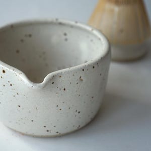 May include: A white ceramic bowl with a speckled brown pattern and a small pouring spout on the rim. The bowl has a rounded shape and a smooth finish. In the background, a blurred light brown bamboo whisk is partially visible out of focus.