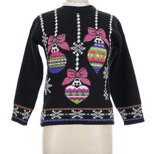 May include: Black sweater with Christmas ornament designs. The sweater features three large ornaments with pink bows and colorful patterns. White snowflakes and vertical lines with white accents decorate the sweater. The cuffs and hem have a patterned border.