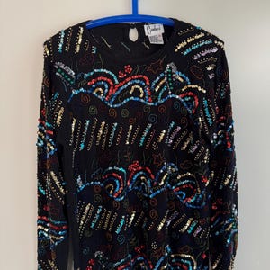 Può includere: Maglione nero a maniche lunghe ornato di paillettes e perline colorate. Il design presenta linee ondulate, stelle e spirali nei toni del rosso, blu, oro e verde. Il maglione è appeso a una gruccia blu.