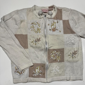 May include: Cream-colored zip-up sweater with embroidered holiday-themed patches. The sweater features squares of cream and tan with embroidered wreaths, candles, and snowflakes. The zipper has a pom-pom detail.