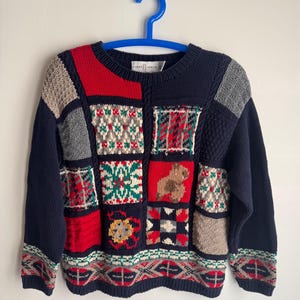 May include: A navy blue knitted sweater with a patchwork design. The sweater features squares of red, grey, and beige with geometric patterns and a teddy bear motif. The label reads "FIRST ISSUE". The sweater has a round neck and long sleeves.