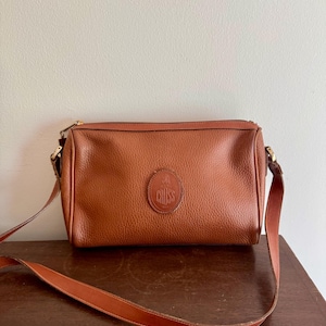 May include: A brown leather crossbody bag with a long adjustable strap. The bag has a rectangular shape with a zipper closure and a circular embossed logo that reads "CRISS". The bag is made of textured leather.