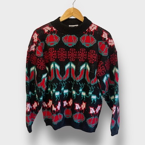 May include: Black knit sweater with a high neckline. The sweater features a repeating pattern of red and green Christmas ornaments, snowflakes, reindeer, and bells. The sweater is hanging on a wooden hanger.