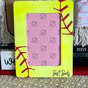 May include: A rectangular neon yellow picture frame featuring red stitching patterns that mimic a softball. The frame includes the handwritten text "Best Buds" in black ink at the bottom right corner and contains a pink insert with repeating logos.