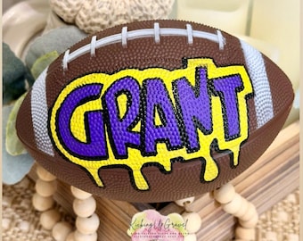 Könnte beinhalten: Ein brauner Mini-Football mit einer Länge von etwa 15 bis 18 cm. Der Football hat weiße Schnürsenkel und ein individuelles Design mit dem Namen GRANT in violetten Buchstaben mit einem gelben Tropfeffekt.