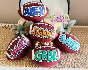 May include: Five brown textured footballs with white laces, each featuring a hand-painted name in a distinct colour. The names ALEX, JOHNNY, JAMES, MILO, and GABE are displayed in bold, block-style lettering with white outlines on the surface of each ball.