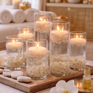 May include: A spa-themed display featuring lit candles in clear glass cylinders. The candles float atop water and are surrounded by decorative beads. The arrangement sits on a wooden tray with smooth stones and a small bottle of oil.