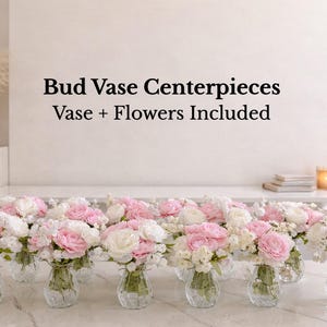 May include: A collection of clear glass bud vases filled with artificial pink and white flowers. The text Bud Vase Centerpieces Vase + Flowers Included is displayed above the vases. Each vase contains a small bouquet of roses and greenery.