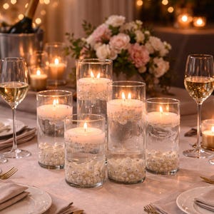May include: A romantic table setting with lit candles in glass cylinders filled with water and pearls. Wine glasses, a bouquet of flowers, and a champagne bottle in a bucket complete the elegant decor. Soft lighting creates a warm ambiance.
