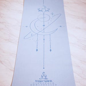Pu&ograve; includere: Un tappetino da yoga azzurro con un design celeste in un blu pi&ugrave; chiaro. Il tappetino presenta una luna crescente, stelle e forme geometriche. Il testo "Yoga Spirit, Stretched & Unstressed" &egrave; stampato in basso.