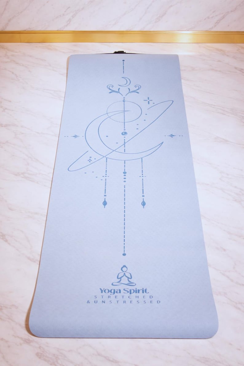 Pu&ograve; includere: Un tappetino da yoga azzurro con un design celeste in un blu pi&ugrave; chiaro. Il tappetino presenta una luna crescente, stelle e forme geometriche. Il testo "Yoga Spirit, Stretched & Unstressed" &egrave; stampato in basso.