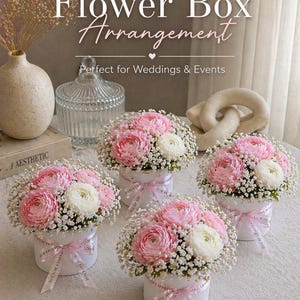 May include: Four white flower boxes each hold a floral arrangement of pink and white flowers, surrounded by small white blossoms. Each box is tied with a pink ribbon. The image includes text that reads "Flower Box Arrangement."