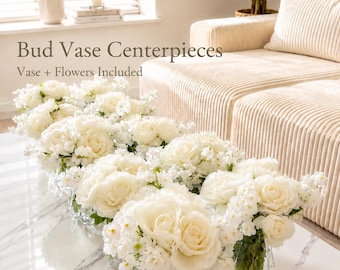 May include: A set of clear glass bud vases with a diamond-cut texture, each filled with white roses and small white filler flowers. The text Bud Vase Centerpieces and Vase + Flowers Included is displayed in the upper left corner of the image.
