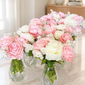May include: A collection of clear, diamond-patterned glass vases, each filled with a lush arrangement of soft pink and white peony-like flowers and small white filler blossoms, featuring green stems visible through the transparent glass.