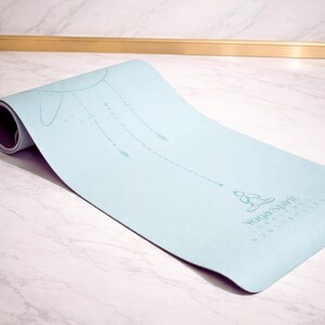Pu&ograve; includere: Un tappetino da yoga azzurro parzialmente arrotolato. Presenta un disegno stampato di un sole e linee con frecce. Le parole "Yoga Spirit" sono stampate. Il tappetino &egrave; su una superficie di marmo bianco.