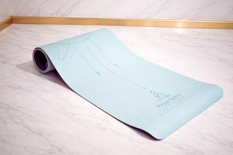 Pu&ograve; includere: Un tappetino da yoga azzurro parzialmente arrotolato. Presenta un disegno stampato di un sole e linee con frecce. Le parole "Yoga Spirit" sono stampate. Il tappetino &egrave; su una superficie di marmo bianco.