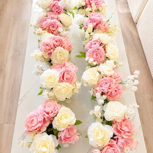 May include: Two rows of floral arrangements featuring a mix of cream and pink roses, interspersed with small white blossoms and green leaves. Each arrangement is composed of multiple blooming rose heads in varying shades of soft pink and cream.