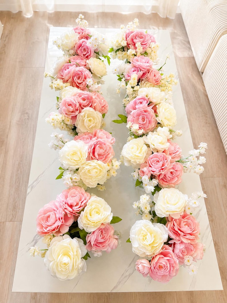 May include: Two rows of floral arrangements featuring a mix of cream and pink roses, interspersed with small white blossoms and green leaves. Each arrangement is composed of multiple blooming rose heads in varying shades of soft pink and cream.