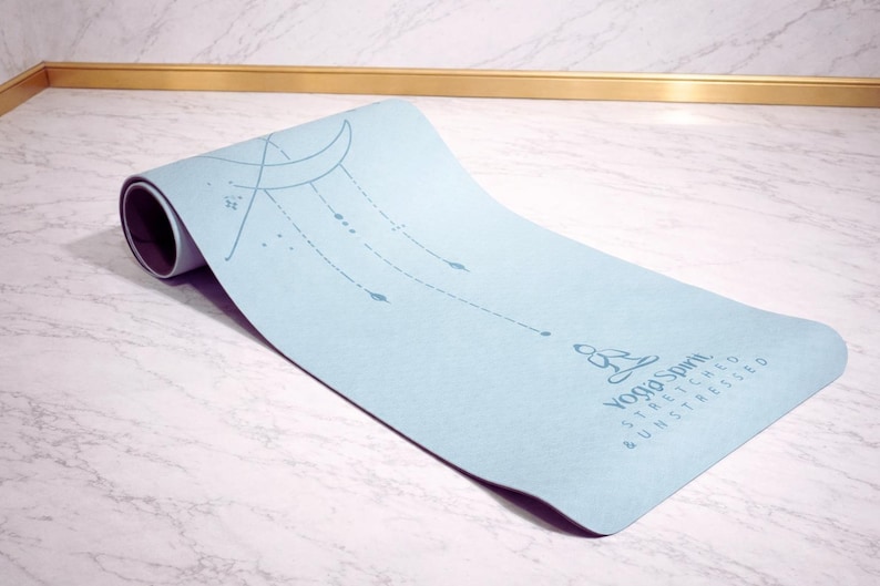 Pu&ograve; includere: Un tappetino da yoga azzurro con un'estremit&agrave; arrotolata. Il tappetino presenta un design a mezzaluna e stelle, insieme alle parole "Yoga Spirit Stretched & Unstressed". Il tappetino &egrave; su una superficie di marmo.