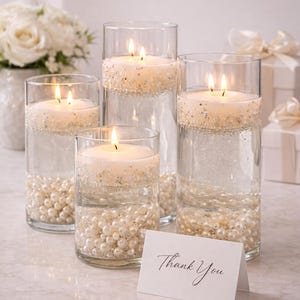 May include: Four clear glass cylinders with lit candles, surrounded by pearls and glitter. The candles float above the pearls, creating a decorative display. A small card with "Thank You" is in front of the cylinders. White flowers and gifts are in the background.