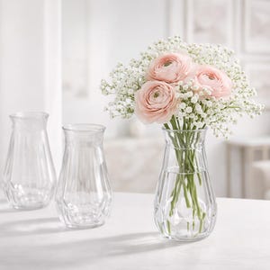 May include: Three clear glass vases with a vertical ribbed pattern and a flared rim. One vase is filled with a bouquet of light pink ranunculus flowers and white baby's breath, while the other two vases remain empty and transparent.
