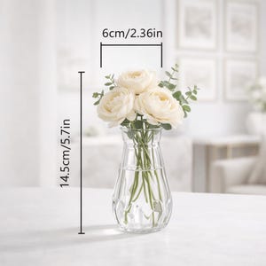 May include: A clear glass vase with a faceted base containing three cream-colored ranunculus flowers and green eucalyptus stems. The vase measures 2.36 inches in width and 5.7 inches in height, as indicated by the visible measurement lines.
