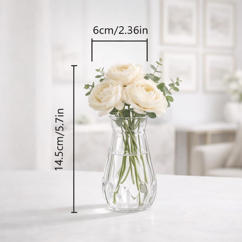 May include: A clear glass vase with a faceted base containing three cream-colored ranunculus flowers and green eucalyptus stems. The vase measures 2.36 inches in width and 5.7 inches in height, as indicated by the visible measurement lines.