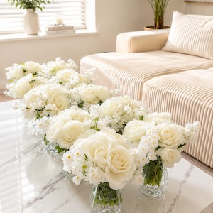 May include: A series of clear, faceted glass vases arranged on a white marble table, each containing a lush bouquet of white roses and small white filler flowers. The stems are visible through the glass, and the blooms are full and rounded.