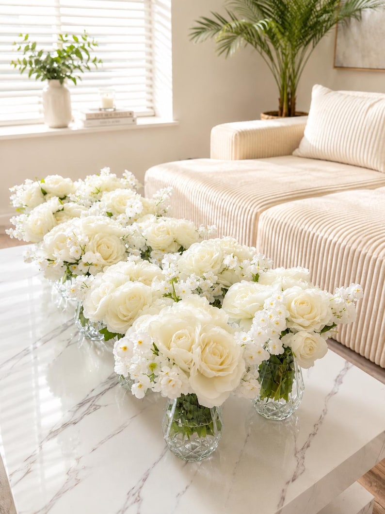 May include: A series of clear, faceted glass vases arranged on a white marble table, each containing a lush bouquet of white roses and small white filler flowers. The stems are visible through the glass, and the blooms are full and rounded.